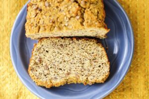 A loaf of gluten free banana bread on a blue plate with one slice cut off, showing a textured interior with visible nuts. The plate is on a yellow textured surface.