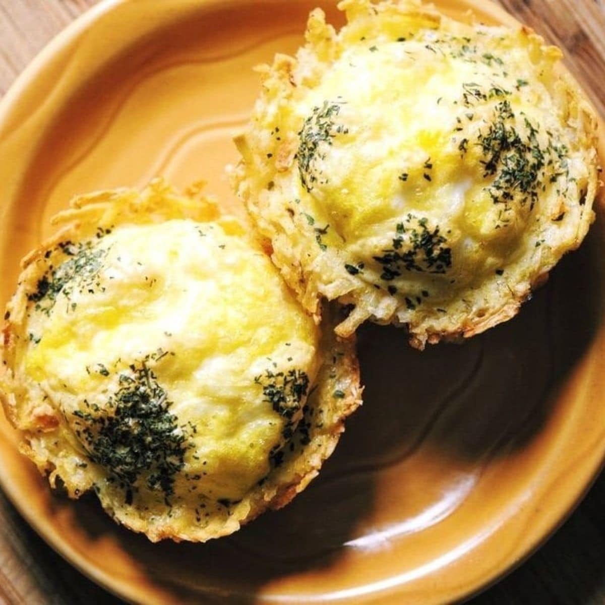 Two savory hash brown egg nests topped with herbs, resting on a round, orange plate.