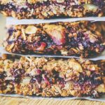 A stack of three granola bars made with oats, nuts, and visible blueberries, separated by pieces of parchment paper on a wooden surface, sits beside a few turmeric crackers gluten-free options for those seeking variety.