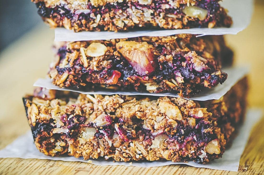 A stack of three granola bars made with oats, nuts, and visible blueberries, separated by pieces of parchment paper on a wooden surface, sits beside a few turmeric crackers gluten-free options for those seeking variety.
