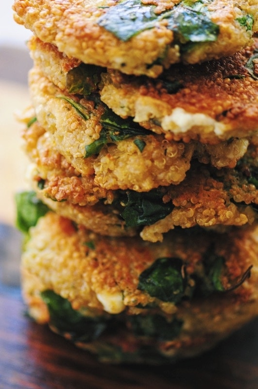 Close-up of a stack of golden brown, baked falafel patties with visible quinoa, leafy greens, and hints of carrot falafel on a wooden surface.