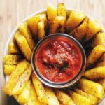 a top view of a white bowl filled with polenta fries with a small jar of marinara in the center