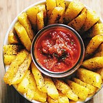 Crispy polenta fries are arranged around a small dish of chunky tomato dipping sauce on a rustic wooden surface.
