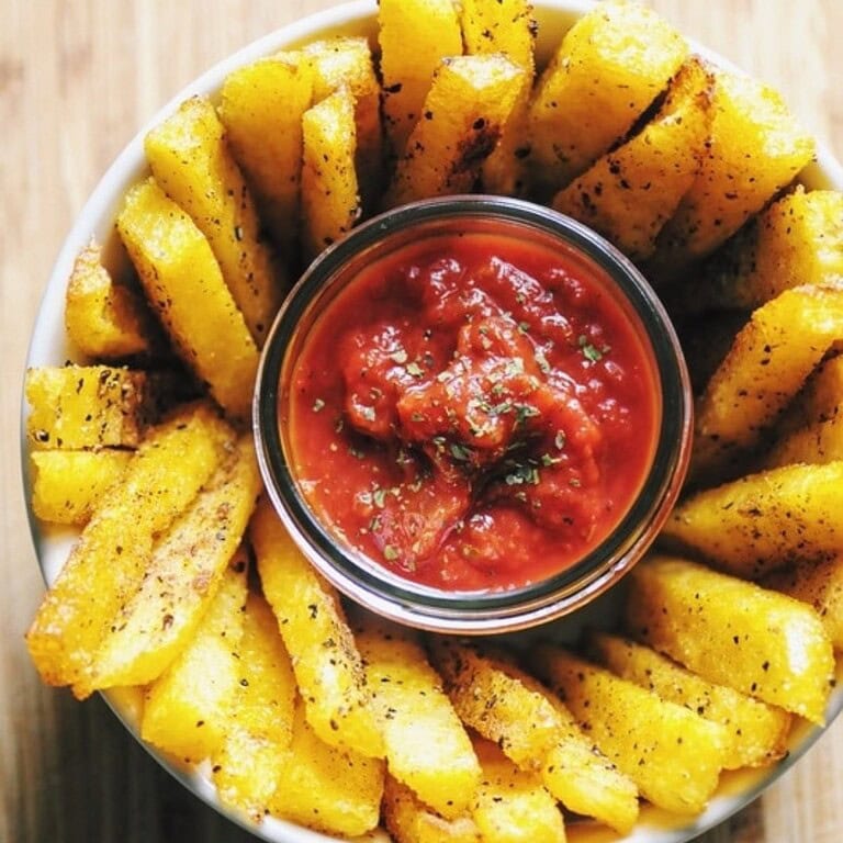 Crispy polenta fries are arranged around a small dish of chunky tomato dipping sauce on a rustic wooden surface.