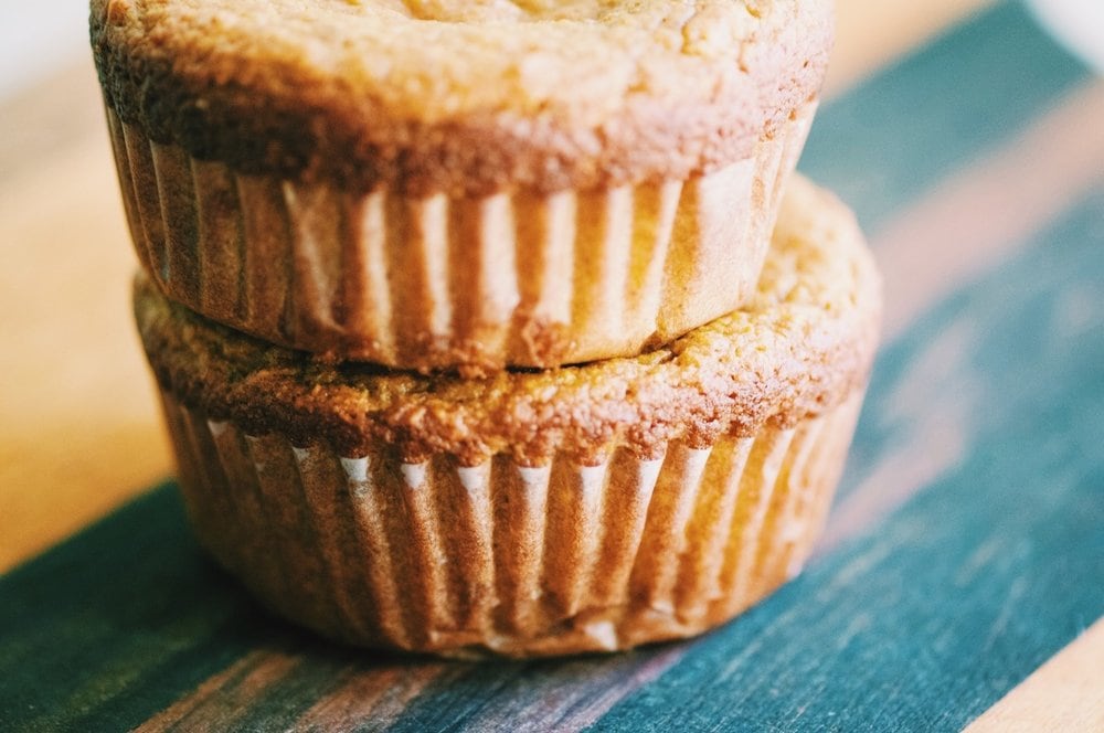 Two muffins in muffin wrappers resting on a wooden cutting board.