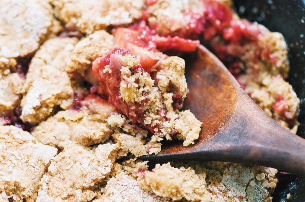 A wooden spoon scoops a raspberry cobbler out of a black pan.