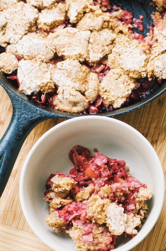 A white bowl sits next to a cast iron skillet, both filled with a cobbler made with pink fruit.