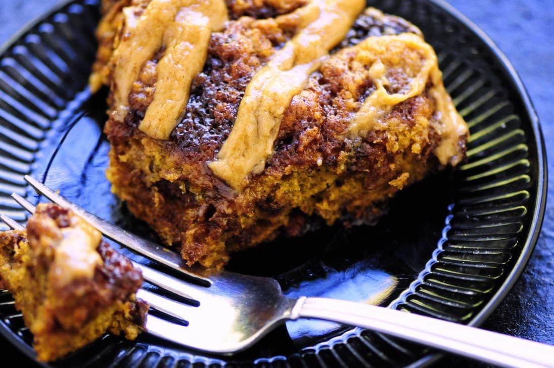 Close-up of a slice of crumbly cake with streaks of frosting on a black plate, accompanied by a fork holding a small piece and a side of vibrant beet hummus.