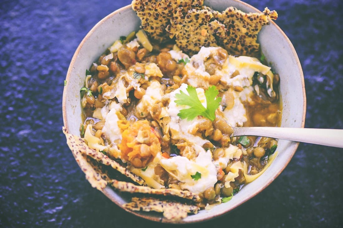A bowl of lentil stew garnished with a dollop of yogurt, parsley, and two pieces of crisp flatbread. A spoon rests inside the bowl, accompanied by a side of BBQ cauliflower bites for an extra savory touch.
