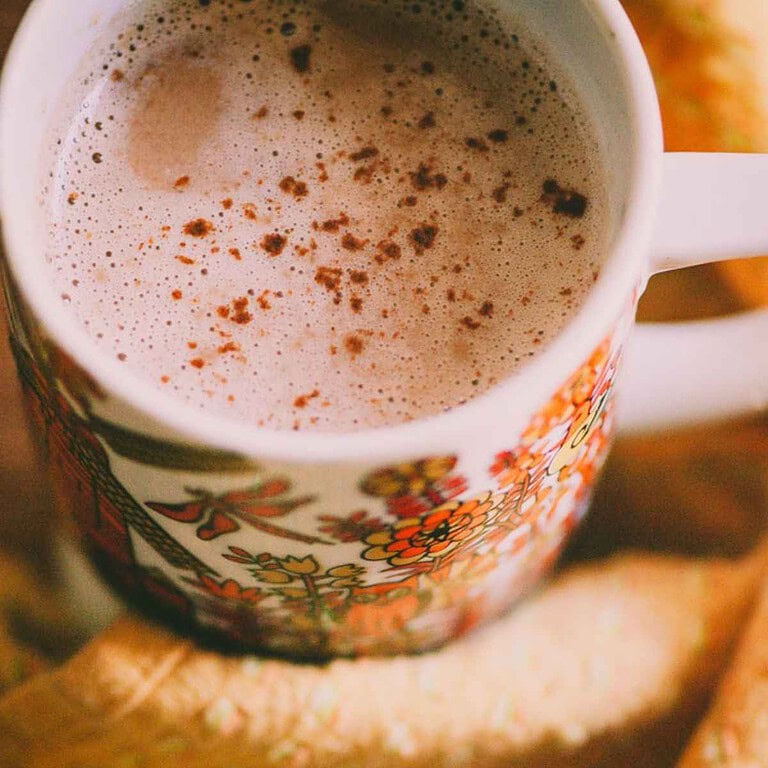 A close-up of a mug filled with frothy tahini hot chocolate, sprinkled with cocoa or cinnamon, in a floral-patterned cup.