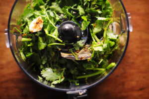 Fresh cilantro, garlic, and spices for chimichurri in a food processor, viewed from above, ready to be blended on a wooden surface.