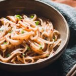 a white bowl filled with sticky noodles on a gray napkin with chopsticks