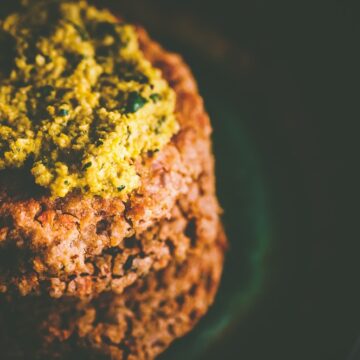 Close-up of a stack of cooked vegan bbq cauliflower patties topped with a greenish-yellow sauce. The patties appear to have a textured surface, and the sauce looks creamy and possibly herb-infused.
