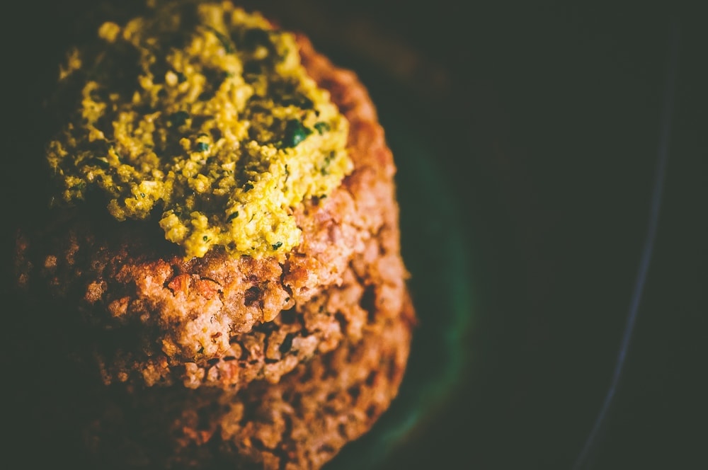 Close-up of a stack of cooked vegan bbq cauliflower patties topped with a greenish-yellow sauce. The patties appear to have a textured surface, and the sauce looks creamy and possibly herb-infused.