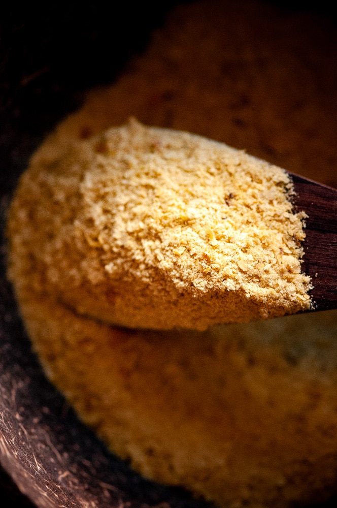 A close up shot of a wooden spoon sticking into a dark wooden bowl of yellow flakes of nutritional yeast.