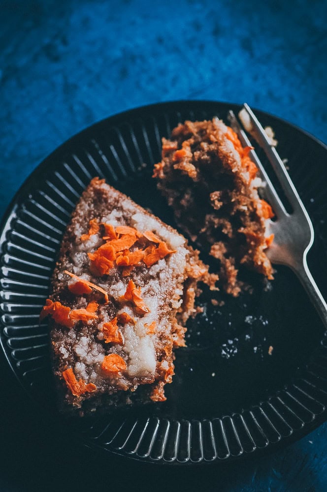A slice of gluten free carrot cake with icing and orange carrot shavings on a black plate, partially eaten, with a fork beside it.