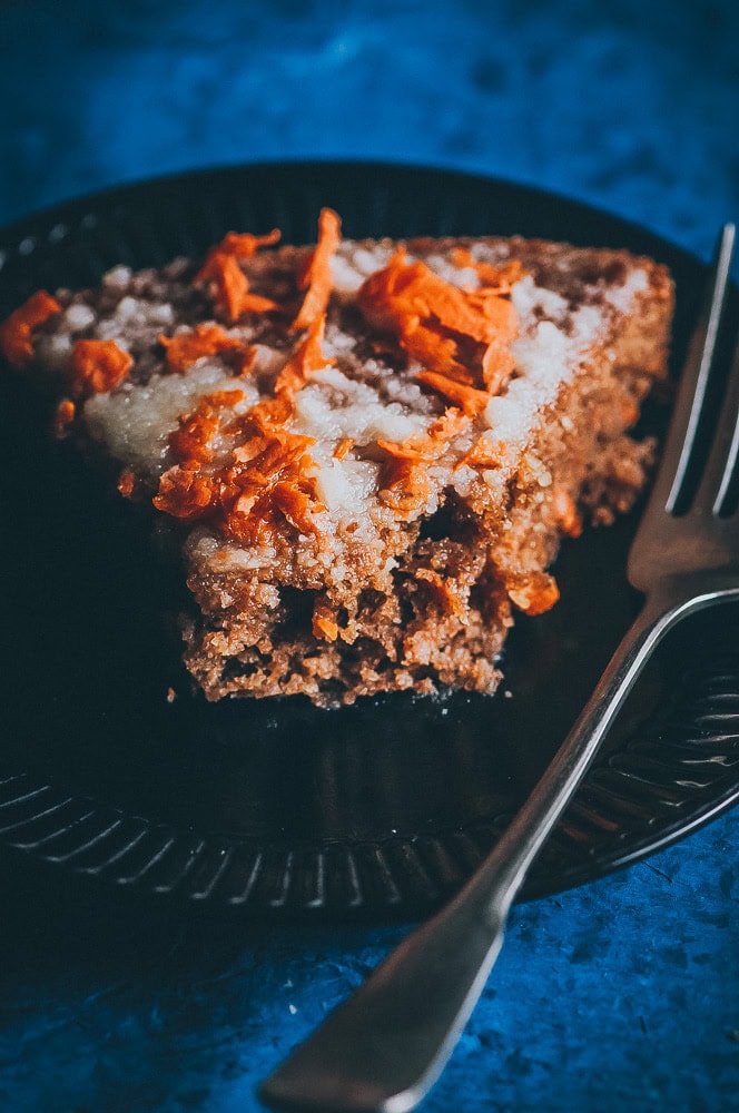 A slice of gluten free carrot cake with grated carrot and frosting on a black plate, next to a fork, on a blue surface.