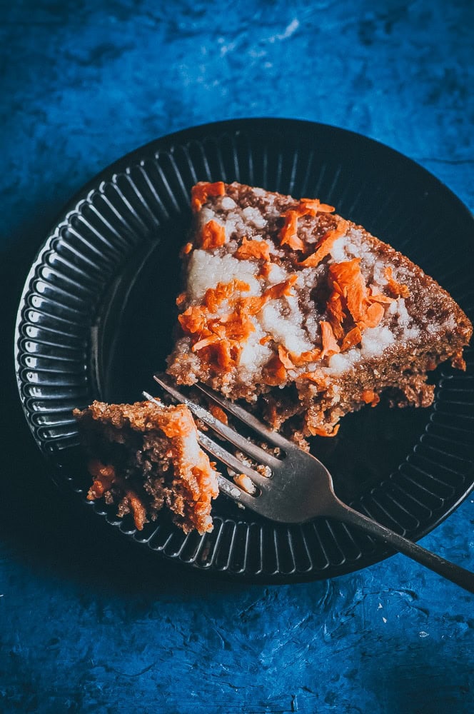 A slice of gluten free carrot cake with icing sits on a black plate, a fork holding a small bite-sized piece beside it.