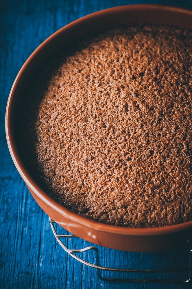 Round gluten free carrot cake in a brown baking pan cooling on a metal rack, viewed from above on a blue wooden surface.
