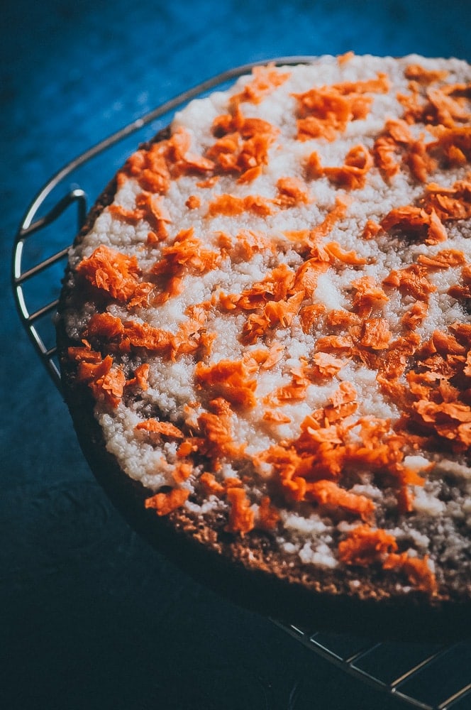 A round gluten free carrot cake with white icing and orange shredded topping sits on a cooling rack against a dark background.