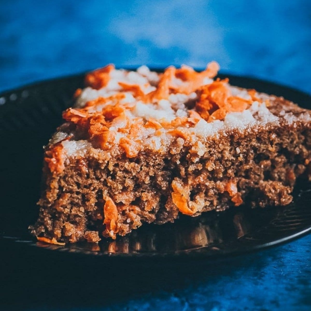 A close-up of a slice of gluten free carrot cake with visible grated carrots and icing on a dark plate against a blue background.