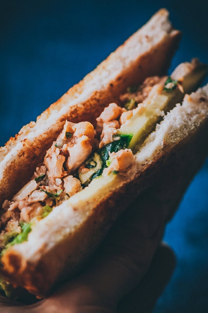 Close-up of a hand holding a delicious chickpea salad sandwich, filled with chopped meat, cucumber slices, and leafy greens between two slices of bread.