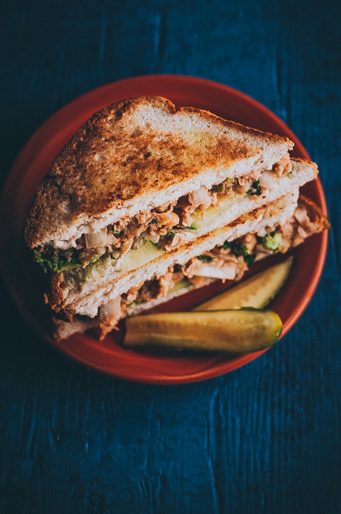 A toasted chickpea salad sandwich filled with meat and vegetables is cut in half and served on a red plate with two pickle spears, set on a dark blue surface.
