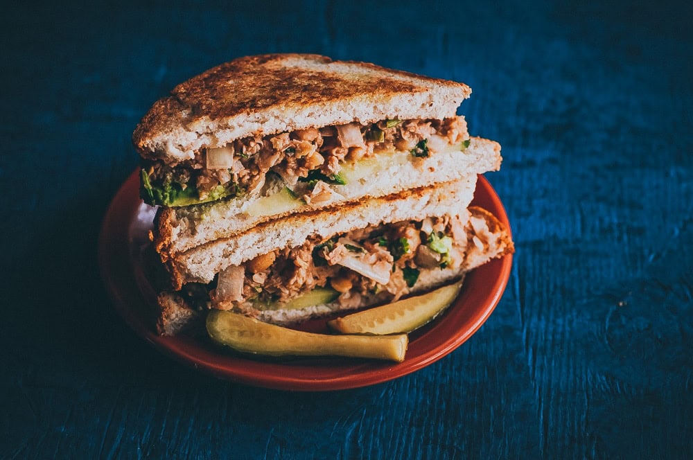 A chickpea salad sandwich cut in half with lettuce and pickles, served on a red plate against a dark blue background.