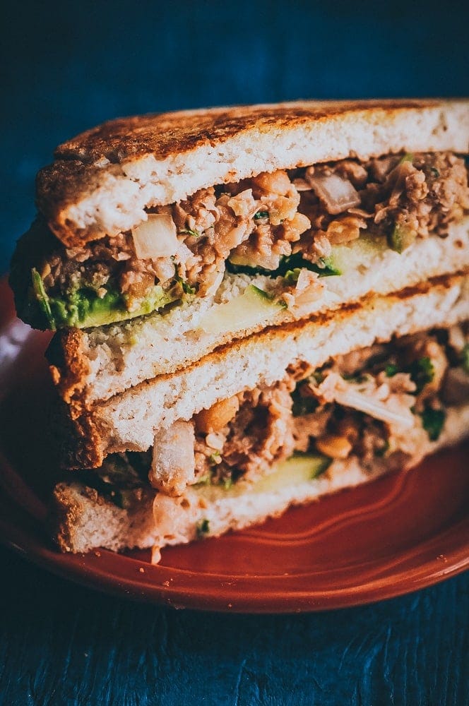 Two halves of a sandwich filled with avocado, chopped onions, and a chickpea salad sandwich mixture are stacked on a red plate.