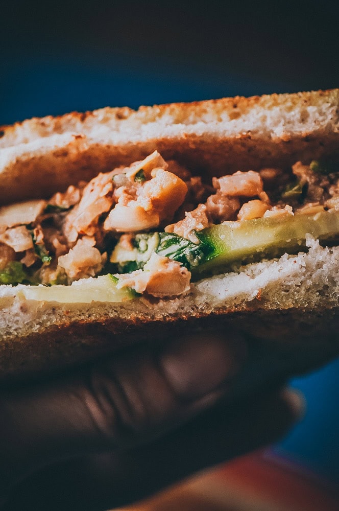 Close-up of a hand holding a chickpea salad sandwich with chopped filling, green vegetables, and thick slices of bread.