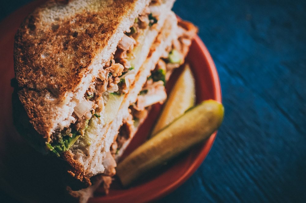 Close-up of a chickpea salad sandwich filled with fresh vegetables on a red plate, served with two pickle spears on the side, set against a dark blue background.