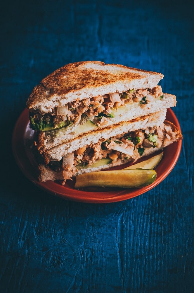 A stacked chickpea salad sandwich cut in half on toasted bread, served on a red plate with a pickle spear on a dark blue wooden surface.