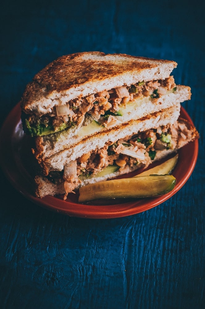 Two halves of a toasted chickpea salad sandwich filled with shredded meat, lettuce, onions, and pickles are stacked on a brown plate with a pickle spear on the side.