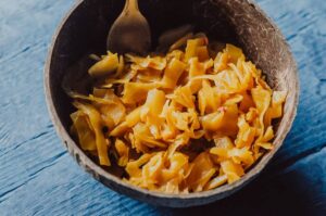 a brown coconut bowl on a blue wooden backdrop filled with yellow turmeric sauerkraut