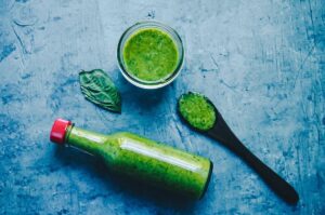 A bottle, glass, and spoon containing green sauce on a blue surface, with a basil leaf nearby, perfect for pairing with vegan zucchini fritters.