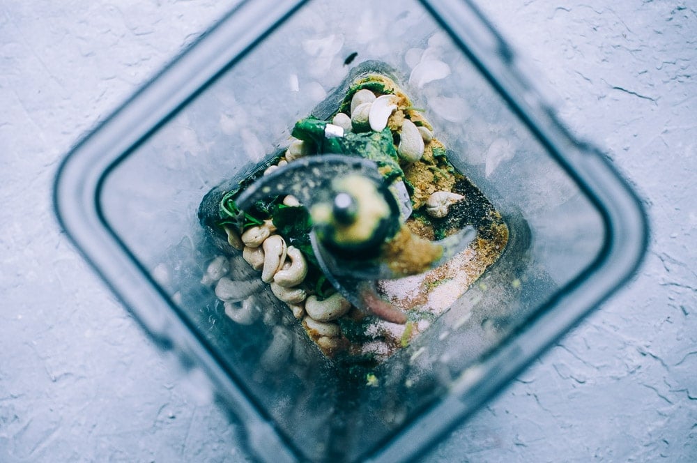 Overhead view of various ingredients, including cashews and greens, in a blender before mixing—perfect for making a fresh vegan cashew pesto—on a light textured surface.
