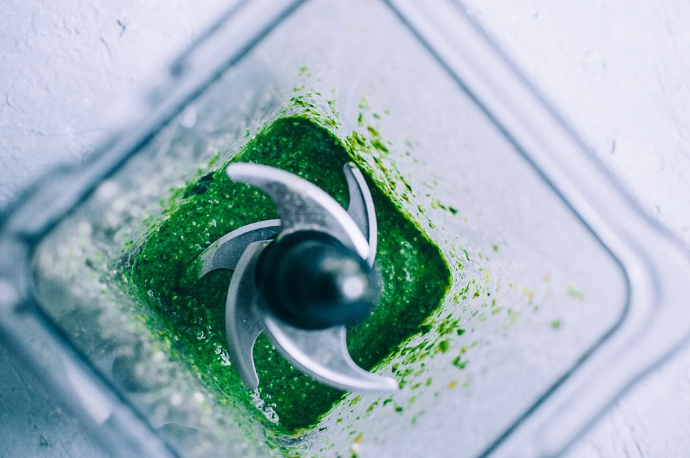 Overhead view of vegan cashew pesto being blended in a blender, with metal blades visible in the center.