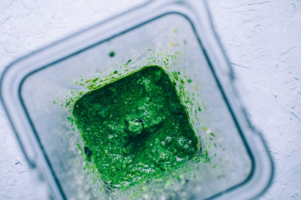 Top-down view of creamy vegan cashew pesto sauce being blended in a blender on a light gray textured surface.