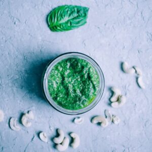 A jar of vegan cashew pesto sits on a gray surface, surrounded by scattered cashew nuts and a single basil leaf.