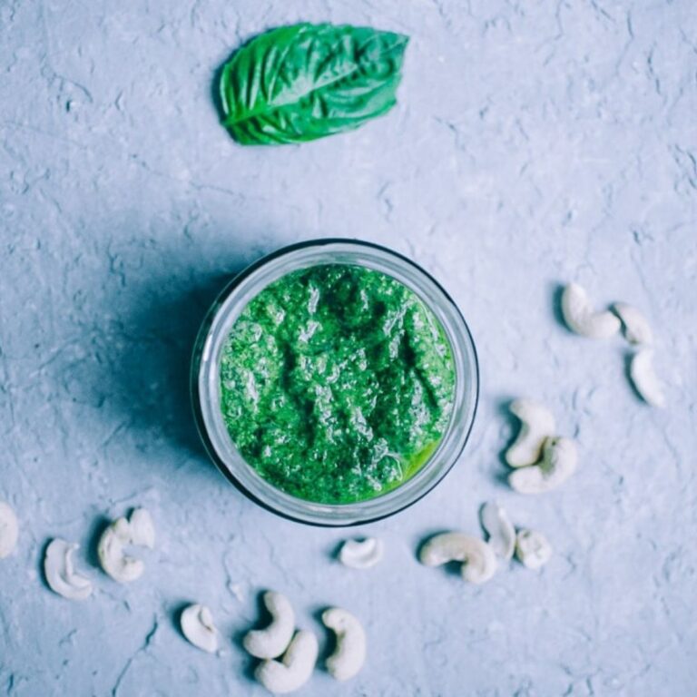 A jar of vegan cashew pesto sits on a gray surface, surrounded by scattered cashew nuts and a single basil leaf.