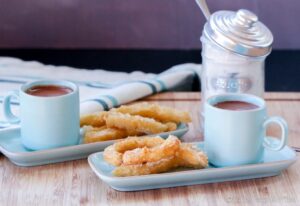 Two blue cups filled with hot chocolate are placed on rectangular plates alongside sugar-coated churros, perfect for dipping. A jar of sugar with a metal lid is in the background, adding a delightful touch to your next tapioca flour recipes endeavor.