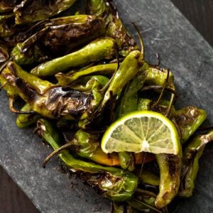 Roasted shishito peppers garnished with a slice of lemon, served on a dark rectangular slate—an ideal choice for those seeking vegetarian keto recipes.