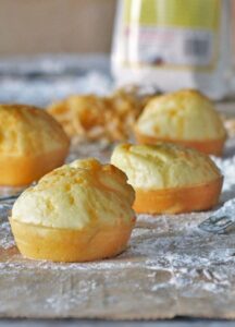 Four freshly baked muffins are placed on a wooden surface dusted with flour. A packet of tapioca flour, perfect for various recipes, is slightly blurred in the background.
