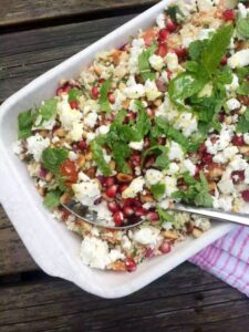 A dish filled with mixed salad ingredients, including crumbled feta cheese, pomegranate seeds, pine nuts, and fresh mint leaves, is served in a white rectangular dish with a spoon. It's one of the delectable vegetarian keto recipes perfect for any meal.