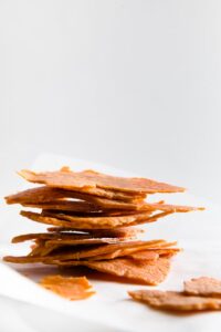 A stack of thin, crispy chicken chips are piled on a white surface against a plain white background, offering a tempting twist for those seeking inspiration beyond vegetarian keto recipes.