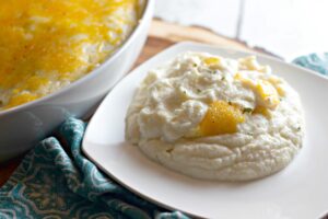 A plate with a serving of mashed cauliflower topped with melted cheese beside a bowl filled with the same dish, placed on a patterned cloth.