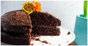 A chocolate cake with a slice removed, decorated with two vibrant yellow and orange flowers, sits on a white plate next to a teal pot containing green plant leaves. It's perfect for those exploring unique tapioca flour recipes.