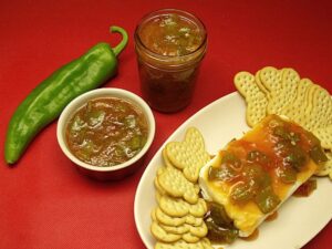 A plate with a cheese-topped block and salsa, surrounded by crackers. A small bowl and a jar filled with salsa, along with a whole green chili pepper, are placed beside the plate on a red surface—perfect for those exploring vegetarian green chile recipes.