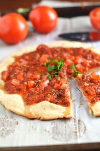 A thin-crust pizza topped with diced tomatoes and herbs, made with a unique touch inspired by tapioca flour recipes. One slice is cut out, revealing the flavorful blend. Fresh tomatoes and knives are visible in the background.