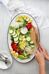 A person arranges wooden salad servers on a platter of salad with sliced avocado, hard-boiled eggs, cucumbers, tomatoes, and a bowl of dressing on a white wooden table with a white cloth nearby, perfect for those seeking delicious vegetarian keto recipes.